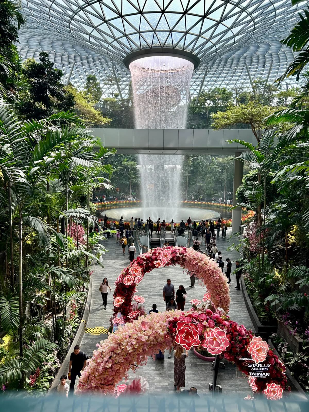The Rain Vortex at Jewel Changi framed by a pink floral arch installation on the garden walkway